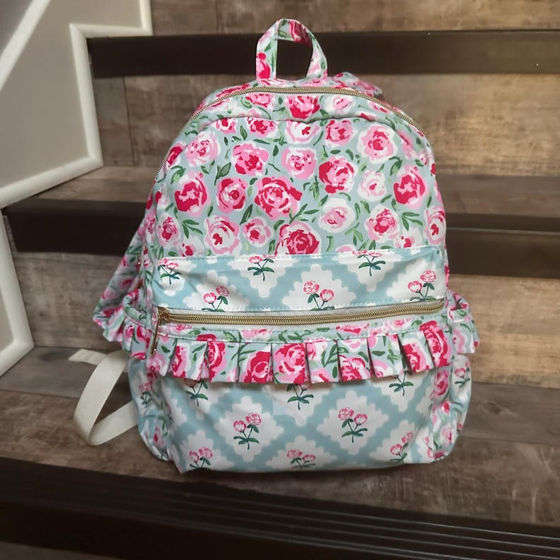May include: A floral backpack with a light blue background and pink roses. The backpack has a top handle, two zippered compartments, and white shoulder straps. Ruffled trim and gold zippers add detail.
