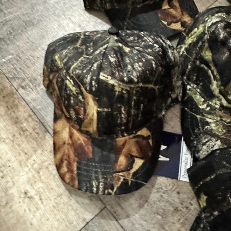 May include: A camouflage baseball cap with a curved brim. The cap features a pattern of brown, green, and tan leaves and branches. The cap is made of a durable fabric and has a black button on top.