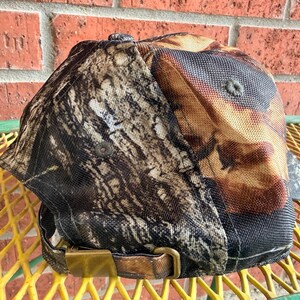 May include: A baseball cap featuring a detailed brown, black, and tan tree bark camouflage pattern. The cap includes a structured crown with a button at the top and an adjustable strap at the back secured with a metallic brass-toned buckle.