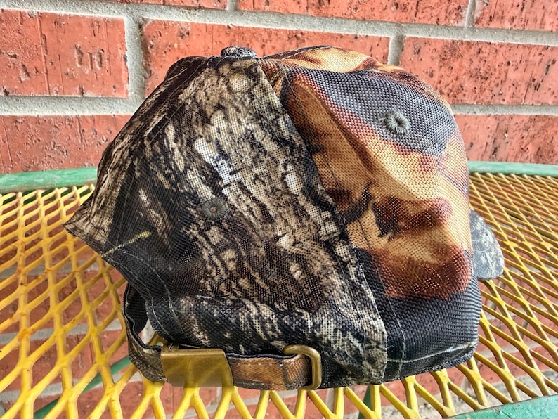 May include: A baseball cap featuring a detailed brown, black, and tan tree bark camouflage pattern. The cap includes a structured crown with a button at the top and an adjustable strap at the back secured with a metallic brass-toned buckle.
