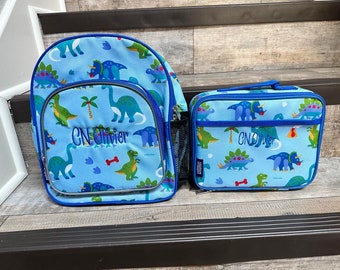 May include: A light blue backpack and a matching rectangular lunch bag, both featuring a colorful dinosaur pattern and the embroidered name "CN Olivier". The backpack includes a front zippered pocket, side mesh pockets, and blue trim.