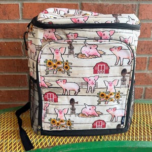 Puede incluir: Una bolsa nevera de color crema con un estampado repetido de cerdos rosas, graneros rojos y girasoles sobre un fondo blanco con vetas de madera. La bolsa tiene un bolsillo lateral de malla negra y una correa para el hombro negra. La bolsa est&aacute; sobre una mesa de metal amarilla.
