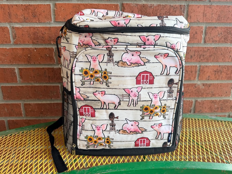 Puede incluir: Una bolsa nevera de color crema con un estampado repetido de cerdos rosas, graneros rojos y girasoles sobre un fondo blanco con vetas de madera. La bolsa tiene un bolsillo lateral de malla negra y una correa para el hombro negra. La bolsa est&aacute; sobre una mesa de metal amarilla.