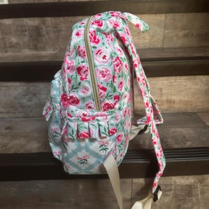 May include: A floral-patterned backpack with a light blue background and pink roses. The backpack has a gold zipper, ruffled pockets, and adjustable straps. The design includes a light blue and white floral pattern on the pockets.