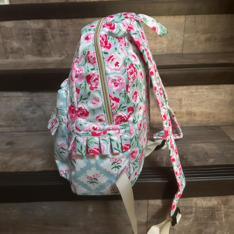 May include: A floral-patterned backpack with a light blue background and pink roses. The backpack has a gold zipper, ruffled pockets, and adjustable straps. The design includes a light blue and white floral pattern on the pockets.