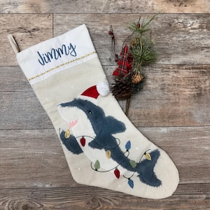 May include: A beige Christmas stocking with a blue shark wearing a Santa hat and holding Christmas lights. The stocking has the name "Jimmy" embroidered on the top.