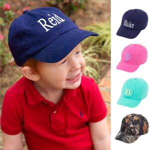 May include: A child wearing a red polo shirt and a navy blue baseball cap with the name "Reid" embroidered in white. The image also shows four different baseball caps with various colours and embroidery, including "Blake", a monogram, and a camouflage pattern.