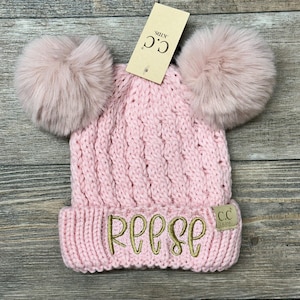 May include: Pink knit beanie with two fluffy pom poms on top and the name "Reese" embroidered in gold on the front. The beanie has a brown tag that says "C.C. Kids."
