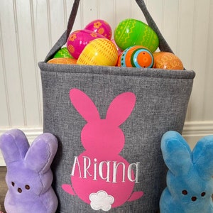 May include: A gray fabric Easter basket with a pink bunny silhouette and the name "Ariana" embroidered on it. The basket is filled with colorful plastic Easter eggs and is surrounded by pastel-colored speckled Easter eggs.