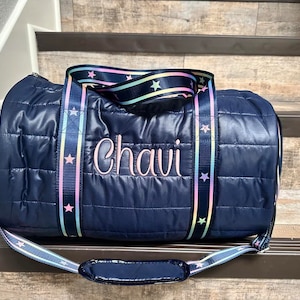 May include: A navy blue quilted duffel bag with a rainbow and star patterned strap. The bag is personalized with the name "Chavi" embroidered in pink.