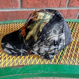 May include: A baseball cap featuring a camouflage pattern in shades of black, white, and brown. The front panel displays a partial orange graphic of a deer antler logo. The cap has a curved brim and a structured crown with visible eyelets.