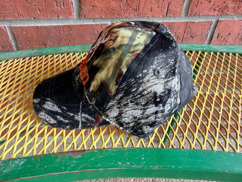 May include: A baseball cap featuring a camouflage pattern in shades of black, white, and brown. The front panel displays a partial orange graphic of a deer antler logo. The cap has a curved brim and a structured crown with visible eyelets.