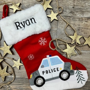 May include: A red Christmas stocking with a white faux fur cuff embroidered with the name Ryan in black. The stocking features white embroidered snowflakes and a police car applique with the word POLICE and a small yellow star on the side.