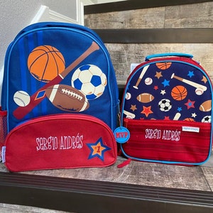 May include: Two red and blue backpacks with a sports theme. The larger backpack has a basketball, baseball, football, and soccer ball on it. The smaller backpack has a repeating pattern of basketballs, baseballs, footballs, soccer balls, and stars. Both backpacks have the name "Sergio Andrés" embroidered on them.