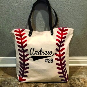 K&ouml;nnte beinhalten: Eine wei&szlig;e Canvas-Tragetasche mit roten Baseballn&auml;hten an den Seiten. Die Tasche hat schwarze Ledergriffe und ein schwarzes Design mit der Aufschrift "Andrew #28".