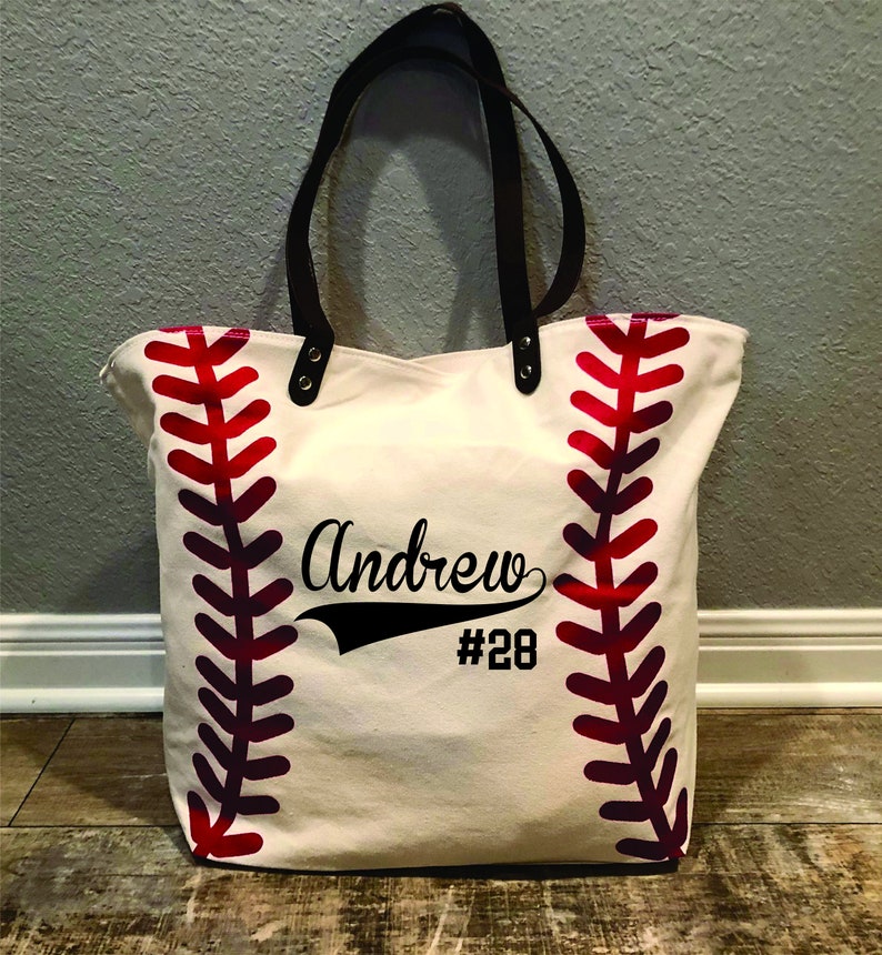 K&ouml;nnte beinhalten: Eine wei&szlig;e Canvas-Tragetasche mit roten Baseballn&auml;hten an den Seiten. Die Tasche hat schwarze Ledergriffe und ein schwarzes Design mit der Aufschrift "Andrew #28".