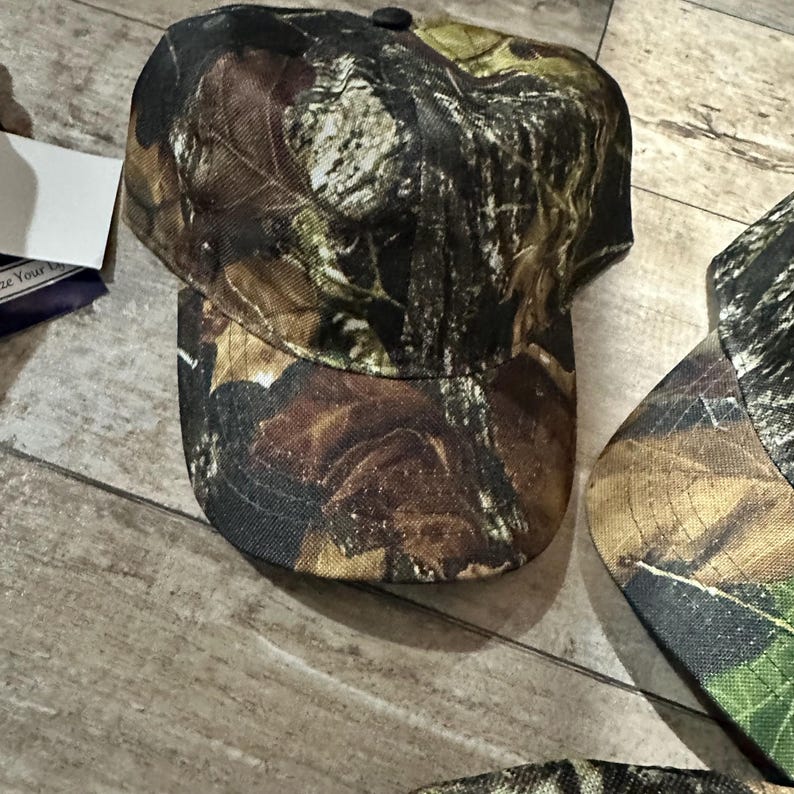 May include: A camouflage baseball cap with a curved brim and a button on top. The cap features a pattern of brown, green, and tan leaves and branches, typical of hunting apparel. The cap is designed to provide shade and protection from the sun.