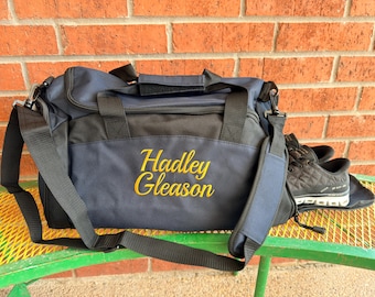 May include: A navy blue and black duffel bag with a black shoulder strap and black handles. The name Hadley Gleason is embroidered in yellow script on the front of the bag. A pair of black athletic sneakers is positioned next to the bag.