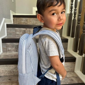 May include: A blue and white striped backpack with navy blue straps. The backpack is worn by a young child. The backpack has a rectangular shape and a zipper closure. The child is wearing a gray t-shirt and navy blue shorts.
