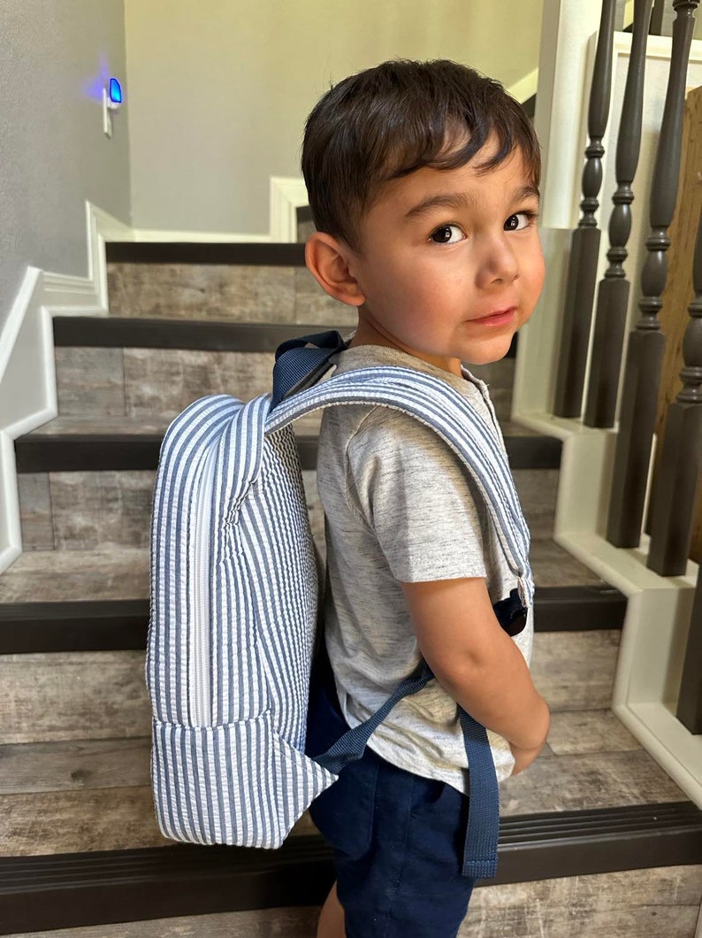 May include: A blue and white striped backpack with navy blue straps. The backpack is worn by a young child. The backpack has a rectangular shape and a zipper closure. The child is wearing a gray t-shirt and navy blue shorts.