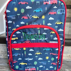 May include: A navy blue rolling suitcase with a red handle and trim. The suitcase features a pattern of various vehicles, including cars, trucks, and airplanes, along with the name "Dawson" embroidered in green. The brand name "stephen joseph" is also visible.