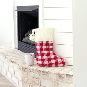 May include: Red and white plaid Christmas stocking with a white cuff and a white pom-pom on the top. The stocking is hanging on a white brick fireplace mantel.
