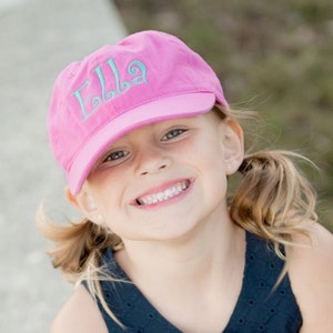 May include: A young girl wearing a pink baseball cap with the name "Ella" embroidered in turquoise on the front. She is smiling and has her hair pulled back in pigtails.
