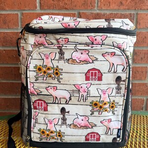 Puede incluir: Una bolsa nevera aislante blanca y negra con estampado de animales de granja. El estampado presenta cerdos rosas, graneros rojos, girasoles y molinos de viento sobre un fondo blanco con un patr&oacute;n de vetas de madera. Tiene un asa negra y un bolsillo lateral de malla.