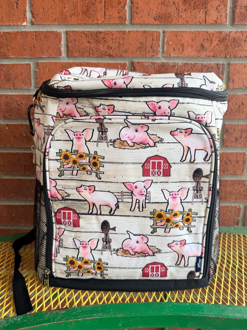 Puede incluir: Una bolsa nevera aislante blanca y negra con estampado de animales de granja. El estampado presenta cerdos rosas, graneros rojos, girasoles y molinos de viento sobre un fondo blanco con un patr&oacute;n de vetas de madera. Tiene un asa negra y un bolsillo lateral de malla.