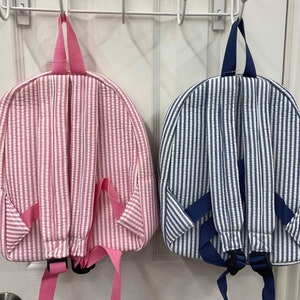 May include: Two seersucker backpacks, one pink and white striped, the other blue and white striped, hang on a white door. Both have padded shoulder straps and a top handle. The pink backpack has pink straps and the blue backpack has navy straps.