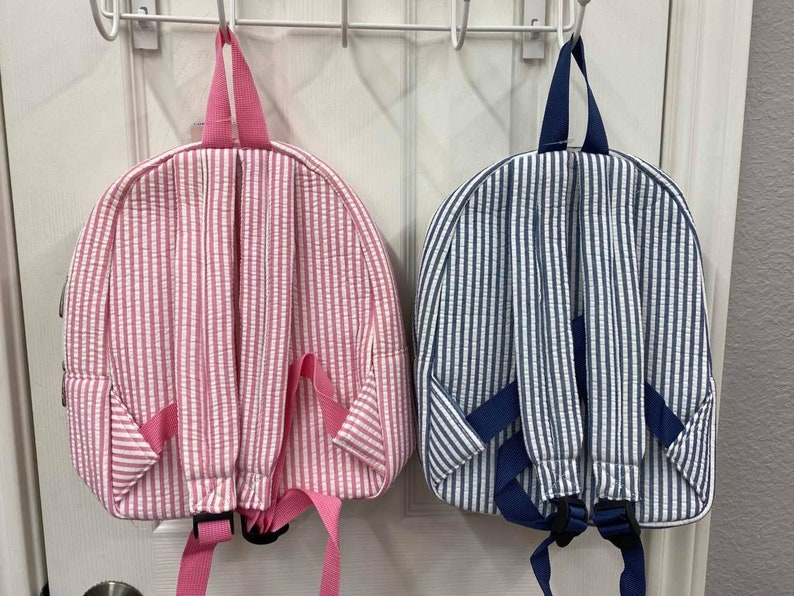 May include: Two seersucker backpacks, one pink and white striped, the other blue and white striped, hang on a white door. Both have padded shoulder straps and a top handle. The pink backpack has pink straps and the blue backpack has navy straps.