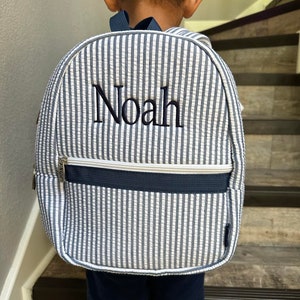 May include: A seersucker backpack with vertical blue and white stripes. The name "Noah" is embroidered in navy blue on the front. The backpack has a navy blue zipper and straps.