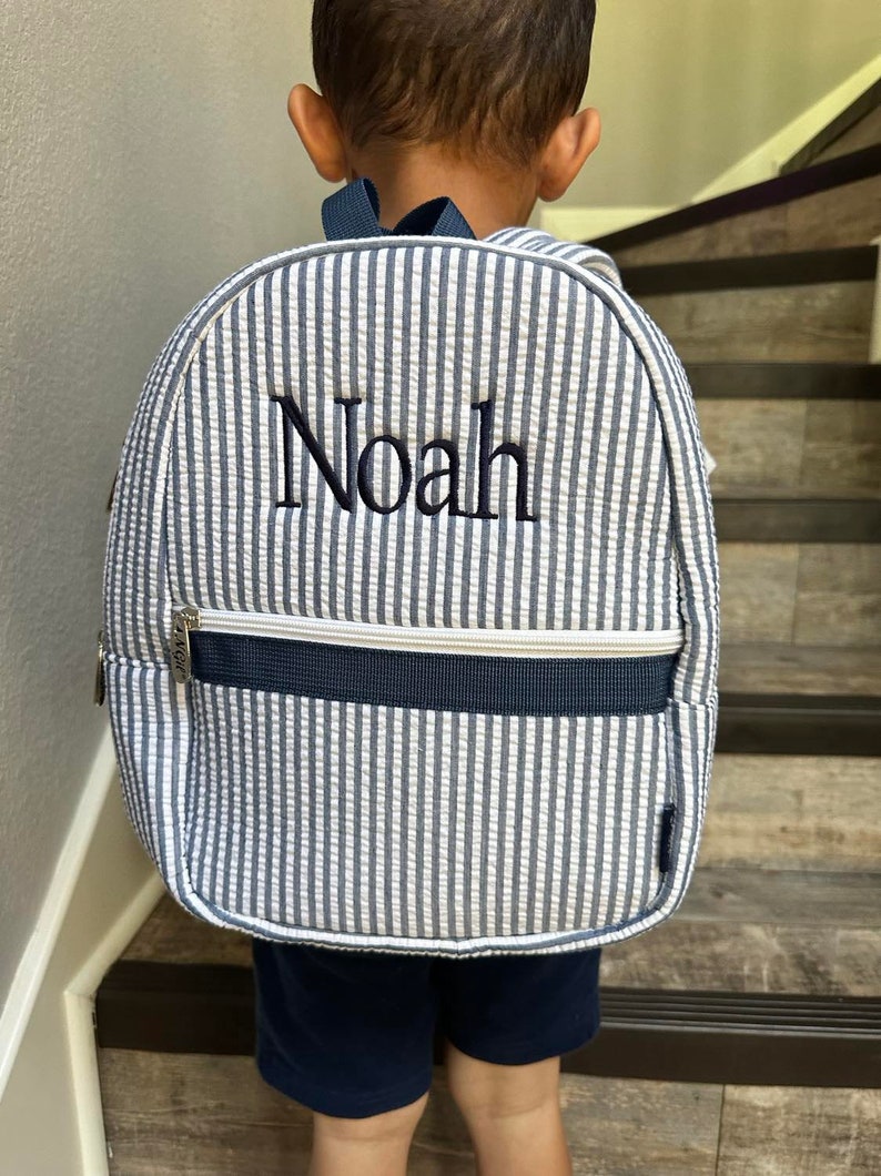 May include: A seersucker backpack with vertical blue and white stripes. The name "Noah" is embroidered in navy blue on the front. The backpack has a navy blue zipper and straps.