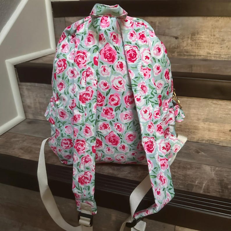May include: A floral backpack with a light blue background and a pattern of pink and red roses. The backpack has white straps and gold-colored hardware. The bag is sitting on wooden stairs.