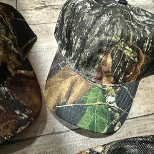 May include: Camouflage baseball cap with a curved brim. The cap features a pattern of green, brown, and black, resembling a forest environment. The cap is made of a durable fabric and has a structured crown. The design is suitable for outdoor activities.