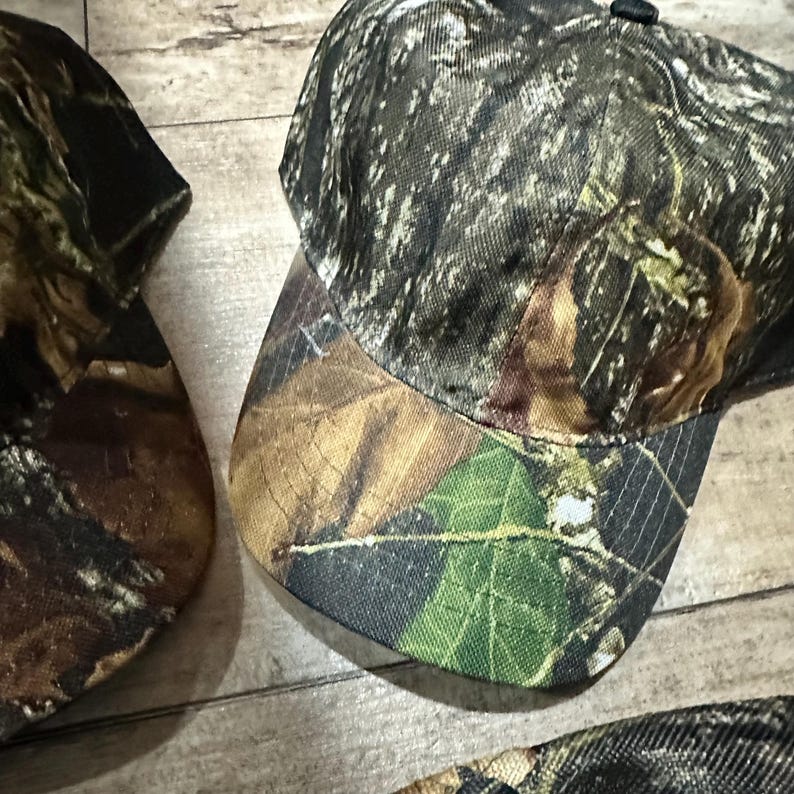 May include: Camouflage baseball cap with a curved brim. The cap features a pattern of green, brown, and black, resembling a forest environment. The cap is made of a durable fabric and has a structured crown. The design is suitable for outdoor activities.