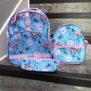 May include: A set of four matching blue backpacks and accessories. The items feature a floral and bee pattern with pink accents. The set includes a large backpack, a smaller backpack, a lunch bag, and a pencil case.