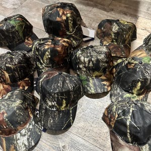 May include: A collection of camouflage baseball caps. The caps feature a pattern of brown, green, and black, designed to blend with natural environments. Each cap has a curved brim and a button on top.