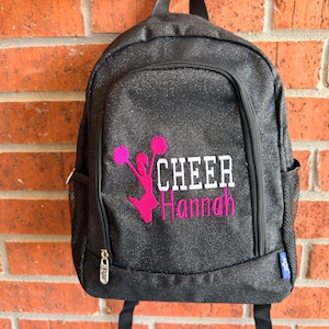 May include: A black glittery backpack featuring a pink embroidered silhouette of a cheerleader holding pom-poms, with the white text CHEER and the pink name Hannah embroidered below. The bag includes a front zippered pocket and side mesh pockets.