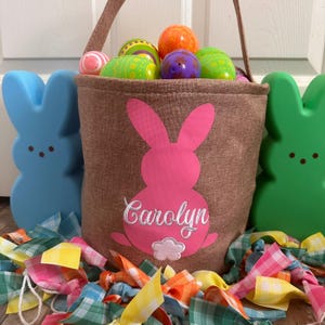 May include: A brown burlap Easter basket with a pink bunny silhouette and the name "Carolyn" embroidered on it. The basket is filled with colorful plastic Easter eggs and is surrounded by colorful plaid ribbons.