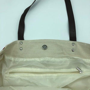 May include: A beige canvas tote bag with brown leather straps and a zippered pocket inside. The bag has a snap closure and metal rivets.