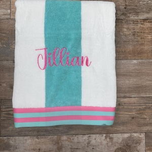 May include: White beach towel with a turquoise stripe down the center and a pink and turquoise border. The name "Jillian" is embroidered in pink script across the turquoise stripe. The towel is folded.
