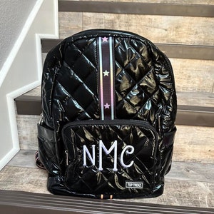 May include: Black quilted backpack with a rainbow star patterned strap and the initials "NMC" embroidered on the front. The backpack has a black zipper and a "TOP TRENDZ" label.