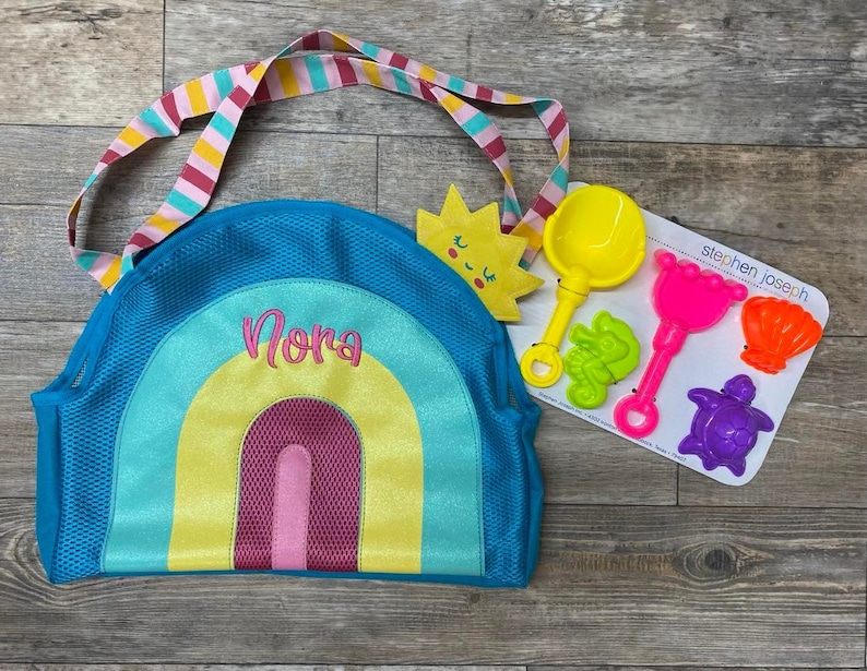 Puede incluir: Una bolsa de playa de malla azul con un dise&ntilde;o de arco&iacute;ris y el nombre "Nora" bordado en ella. La bolsa tiene un sol amarillo con una cara sonriente y un juego de juguetes de playa de pl&aacute;stico de colores unidos a ella.