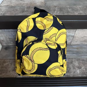 May include: A yellow and black softball-themed backpack. The backpack features a repeating pattern of yellow softballs with red stitching on a navy blue background. It has a small front pocket and a black strap.