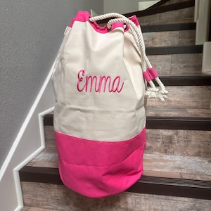 May include: A canvas duffel bag with a cream-colored upper section and a bright pink lower section. The bag features a drawstring closure with a thick white rope and pink accents. The name "Emma" is embroidered in pink script on the front.