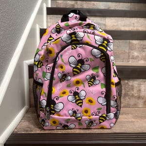 May include: Pink backpack with a bee and sunflower pattern. The backpack has a black zipper and mesh side pockets.