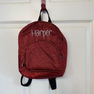 May include: A red glitter backpack with the name "Harper" embroidered in white. The small backpack has a front pocket, black straps, and is hanging on a white door.