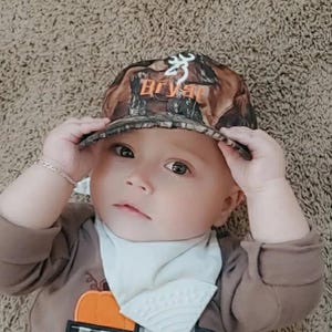 May include: A baby wearing a camouflage baseball cap with the name "Bryant" in orange lettering. The baby is also wearing a brown long-sleeved shirt and a white bib. The background is a light brown carpet.