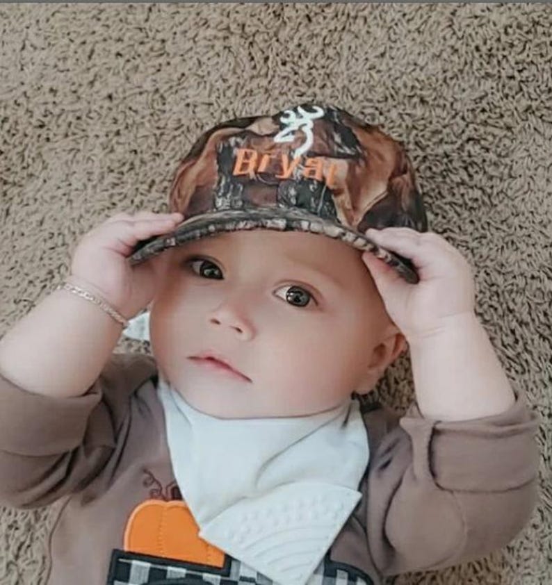 May include: A baby wearing a camouflage baseball cap with the name "Bryant" in orange lettering. The baby is also wearing a brown long-sleeved shirt and a white bib. The background is a light brown carpet.